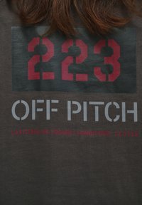 Dark gray t-shirt featuring bold red and gray text: "223 OFF PITCH" with coordinates "LATITUDE: 55.702668 | LONGITUDE: 12.5724."