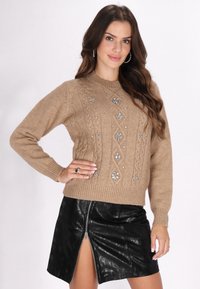 Beige Strickpullover mit dekorativen silbernen Perlen und Zopfmuster, kombiniert mit einem schwarzen Leder-Minirock mit einem Reißverschluss vorne.