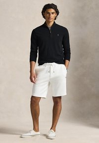 Joven con suéter negro con cremallera hasta el pecho, pantalones cortos blancos con cordón, zapatillas blancas y reloj de pulsera, posando de pie frente a un fondo neutro.