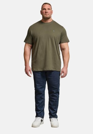 En mand står med front mod kameraet iført en olivengrøn T-shirt, mørkeblå jeans og hvide sneakers med sorte swooshes på en ensfarvet baggrund.