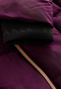 Veste d'hiver violette avec une texture lisse, dotée d'un poignet en ribbed noir et d'une fermeture éclair dorée, affichant un branding proéminent sur le poignet.