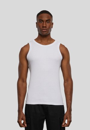 Junger Mann trägt ein ärmelloses weißes geripptes Tanktop und schwarze Hose, steht vor einem einfarbigen grauen Hintergrund.