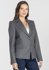 Kobieta w dopasowanej szarej marynarce z czarnymi guzikami, nosząca białą koronkową bluzkę i ciemnoniebieskie dżinsy, stojąca na jednolitym tle.