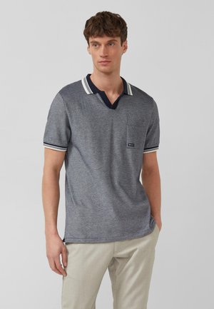 Młody mężczyzna w szarej koszulce polo z krótkim rękawem, granatowym kołnierzem i beżowych spodniach, stojący z jedną ręką w kieszeni.