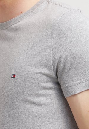 Nahaufnahme einer Person, die ein hellgraues Kurzarm-T-Shirt mit einem kleinen rechteckigen rot-weiß-blauen Logo auf der Brust trägt.