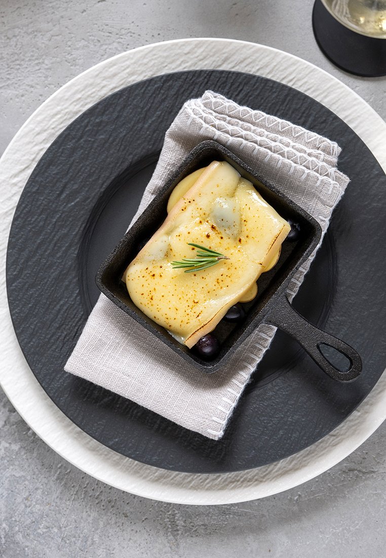 Żeliwne naczynie z roztopionym serem na jagodach, umieszczone na teksturowanym szarym talerzu z białą krawędzią, w towarzystwie złożonego białego serwetki.