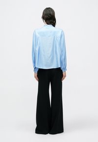 Svetlomodrá košeľa z hladkej látky s dlhými balónovými rukávmi a štruktúrovaným zadným dielom; v kombinácii s čiernymi nohavicami so širokými nohavicami.