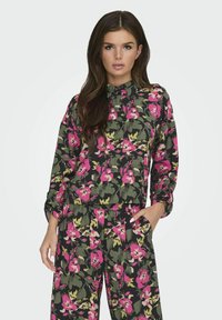 Blomsterpræget, letvægts stofskjorte med en mørk baggrund, der har rosa, gule og grønne blomster. Inkluderer en krave og knaplukning foran.