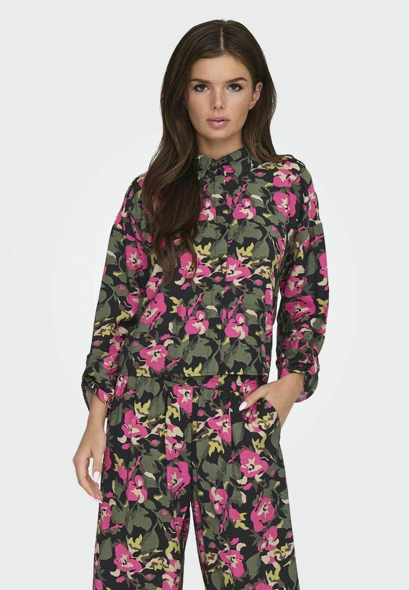 Blomsterpræget, letvægts stofskjorte med en mørk baggrund, der har rosa, gule og grønne blomster. Inkluderer en krave og knaplukning foran.