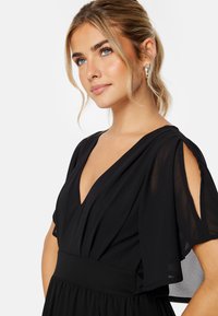 Schwarzes Kleid mit V-Ausschnitt, kurzen Capeärmeln und tailliertem Schnitt. Leichter Stoff mit einem fließenden Rock und subtilen Drapierungsdetails.