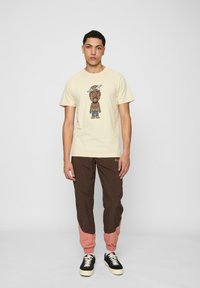 Cream t-shirt with cartoon graphic and short sleeves, paired with brown and pink color-blocked joggers and black sneakers.