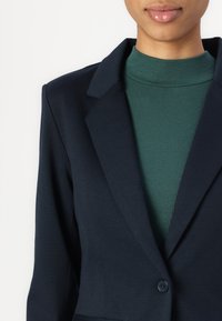 Blazer marine avec revers crantés et fermeture à un bouton, superposé à un col roulé côtelé vert foncé texturé. Tissu lisse.