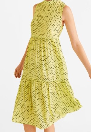 Robe jaune sans manches à pois noirs, col montant, taille cintrée et jupe fluide portée par une personne aux bras légèrement étendus.