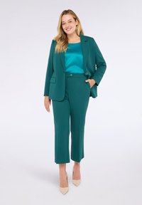 Kobieta uśmiechająca się w tealdowej marynarce i spodniach, z tealdową satynową bluzką i beżowymi szpilkami, stojąca na tle jednolitego, jasnego tła.