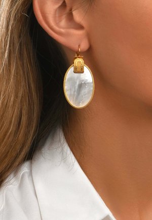 Boucle d'oreille anneau en or avec un grand pendentif ovale blanc en nacre portée à l'oreille d'une femme aux cheveux châtain clair et col de chemise blanc.