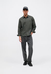 Camisa de manga larga verde oscuro con botonera y bolsillo en el pecho, combinada con vaqueros grises de corte holgado y zapatos negros.