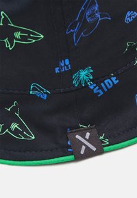 Svart caps med grønne og blå hai-mønstre, teksten "INGEN REGLER" og en grønn kant på skjermen; stoffet ser glatt og holdbart ut.