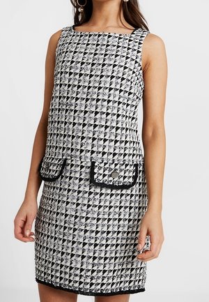 Mouwloze zwart-witte pied-de-poule jurk met voorflapzakken en decoratieve knopen, gedragen door een vrouw tegen een effen achtergrond.