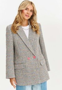 Dvojřadý sako ve stylu houndstooth z texturované látky s barevnými flíčky, růžovými knoflíky a předními kapsami, spárované s bílou košile.