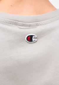 Γκρι ύφασμα με μικρό κεντητό κόκκινο, μαύρο και λευκό λογότυπο "C" στο κέντρο κοντά στη λαιμόκοψη.