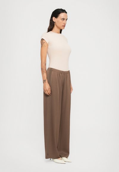 Woman with long dark hair wearing a beige fitted short-sleeve top, loose brown wide-leg pants, white pointed heels, and arm tattoos, standing sideways.