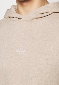 Sudadera color tostado con un sutil bordado blanco de logo de perchero en el pecho. Tejido suave, corte relajado y un diseño básico de capucha.