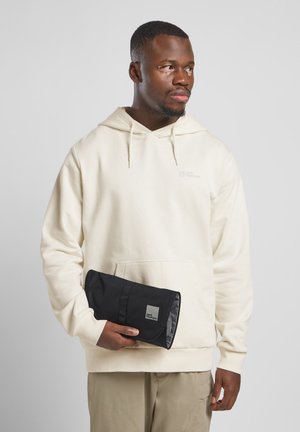 Helle beige Kapuzenjacke mit einer Kängurutasche, kombiniert mit einem schwarzen Beutel, der ein graues Logo und eine glänzende Oberfläche aufweist. Lässige Passform, weicher Stoff.