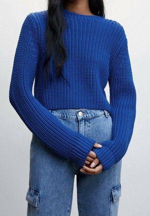 Kvinde iført en lys blå chunky strikket sweater med lange ærmer kombineret med lyseblå cargobukser i høj talje, hænderne foldet foran.