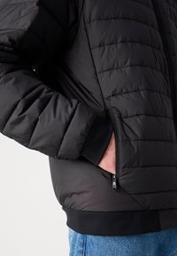 Veste matelassée noire avec texture capitonnée, dotée de poches zippées sur les côtés et de poignets côtelés, vue de la taille vers le haut.