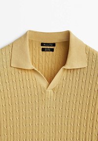 Khaki dzianinowy sweter polo z teksturalnym wzorem w warkocze, dekoltem w kształcie litery V i płaskim kołnierzykiem. Metki wskazują markę oraz instrukcje dotyczące pielęgnacji.