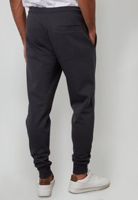 Mand iført sorte joggerbukser og hvide sneakers, stående med hænderne i lommerne, vendt væk mod en ensfarvet baggrund.