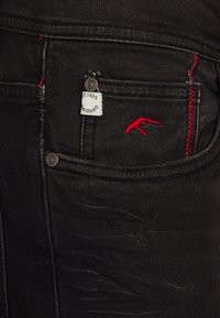 Jean en denim noir avec un logo rouge brodé, une étiquette visible avec "EST. 1993" et des surpiqûres rouges contrastantes sur la poche.
