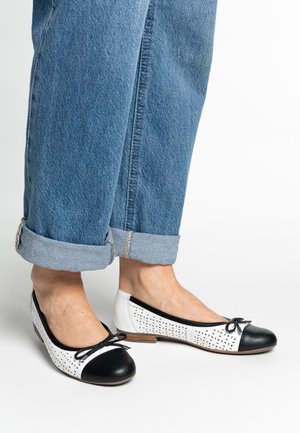 Black and white flat shoes with perforated white leather, rounded black toe cap, and a small bow accent on the front. Worn with rolled denim.