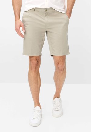 Man draagt beige knielange shorts, witte sneakers en een wit shirt, staat met handen in de zakken op een witte achtergrond.