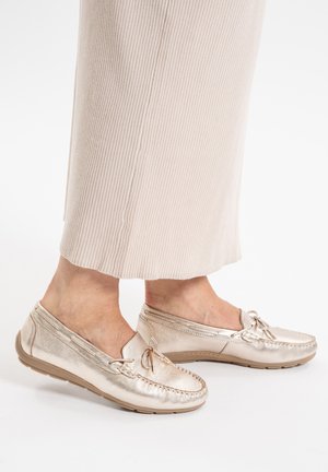 Persona vestida con pantalones beige acanalados y mocasines metálicos dorados con detalle de lazo sobre fondo blanco.