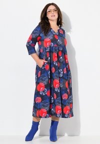 Robe à motif floral bleu foncé avec des accents rouges et bleus. Décolleté en V, manches trois-quarts et poches. Portée avec des bottines à talons bleues.