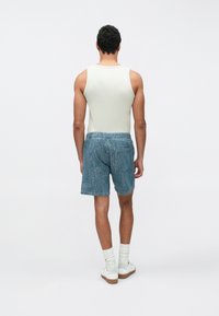 Męska bezrękawnik w jasnym kolorze, zestawiona z niebieskimi szortami dżinsowymi, wyposażonymi w elastyczny pas i tylną kieszeń, na jednolitym tle.