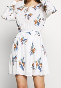 Mujer con un vestido blanco de manga larga, con patrones florales en azul, naranja y verde y una cintura elástica, arreglándose el cabello.