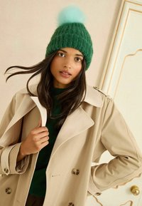 Zelená pletená čepice s světle modrým bambulí, nošená s béžovým dvouřadým kabátem a tmavě zeleným topem, na neutrálním pozadí.