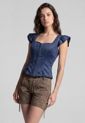 Frau trägt ein eng anliegendes blaues Oberteil mit Rüschenärmeln und braune bestickte Leder-Shorts, steht vor einem schlichten grauen Hintergrund.
