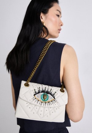 Femme aux longs cheveux noirs portant un haut sans manches bleu marine et un sac matelassé blanc avec un motif d'œil brodé et une bandoulière en chaîne dorée sur l'épaule.