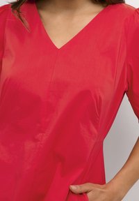 Nainen, jolla on päällään kirkkaanpunainen V-pä�äntie-mekko lyhyillä hihoilla, hänen oikea kätensä lepää sivutaskussa.