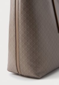 Beež käsipott, millel on tekstureeritud pind ja omavahel seotud muster. Siledad nahkaktsendid ääres ja lukul.