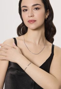 Srebrno-kolorowy naszyjnik i bransoletka na kobiecie noszącej czarną satynową bluzkę. Biżuteria ma wydłużone ogniwa o wypolerowanej powierzchni.