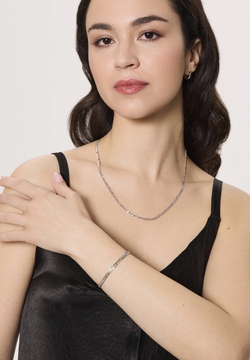 Srebrno-kolorowy naszyjnik i bransoletka na kobiecie noszącej czarną satynową bluzkę. Biżuteria ma wydłużone ogniwa o wypolerowanej powierzchni.