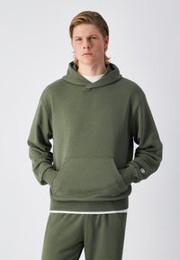 Sudadera color verde oliva hecha de tela suave, con un bolsillo frontal, capucha ajustable con cordón y puños acanalados. Combinada con pantalones a juego.