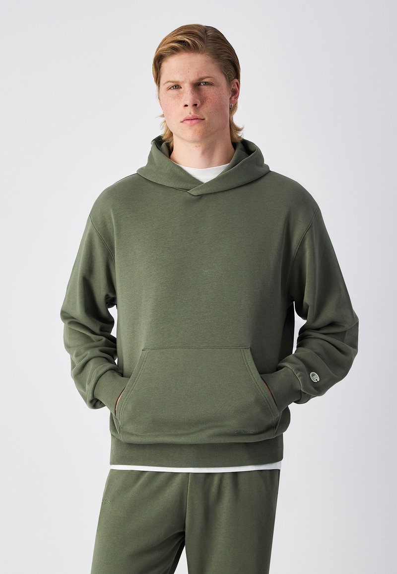 Sudadera color verde oliva hecha de tela suave, con un bolsillo frontal, capucha ajustable con cordón y puños acanalados. Combinada con pantalones a juego.