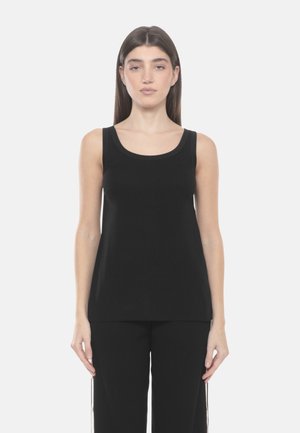 Giovane donna con lunghi capelli castani, che indossa un top nero senza maniche e pantaloni neri, in piedi contro uno sfondo bianco uniforme.