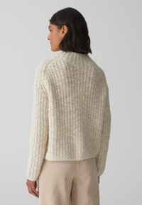 Maglione spesso in crema con una texture a coste, caratterizzato da un collo alto e spalle scese. Indossato con pantaloni in corduroy beige.