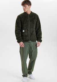 Fleece bunda v tmavě zelené barvě se zipem vpředu, kapsou na hrudi a žebrovanými manžetami. V kombinaci s olivovými cargo kalhotami a bílými teniskami.